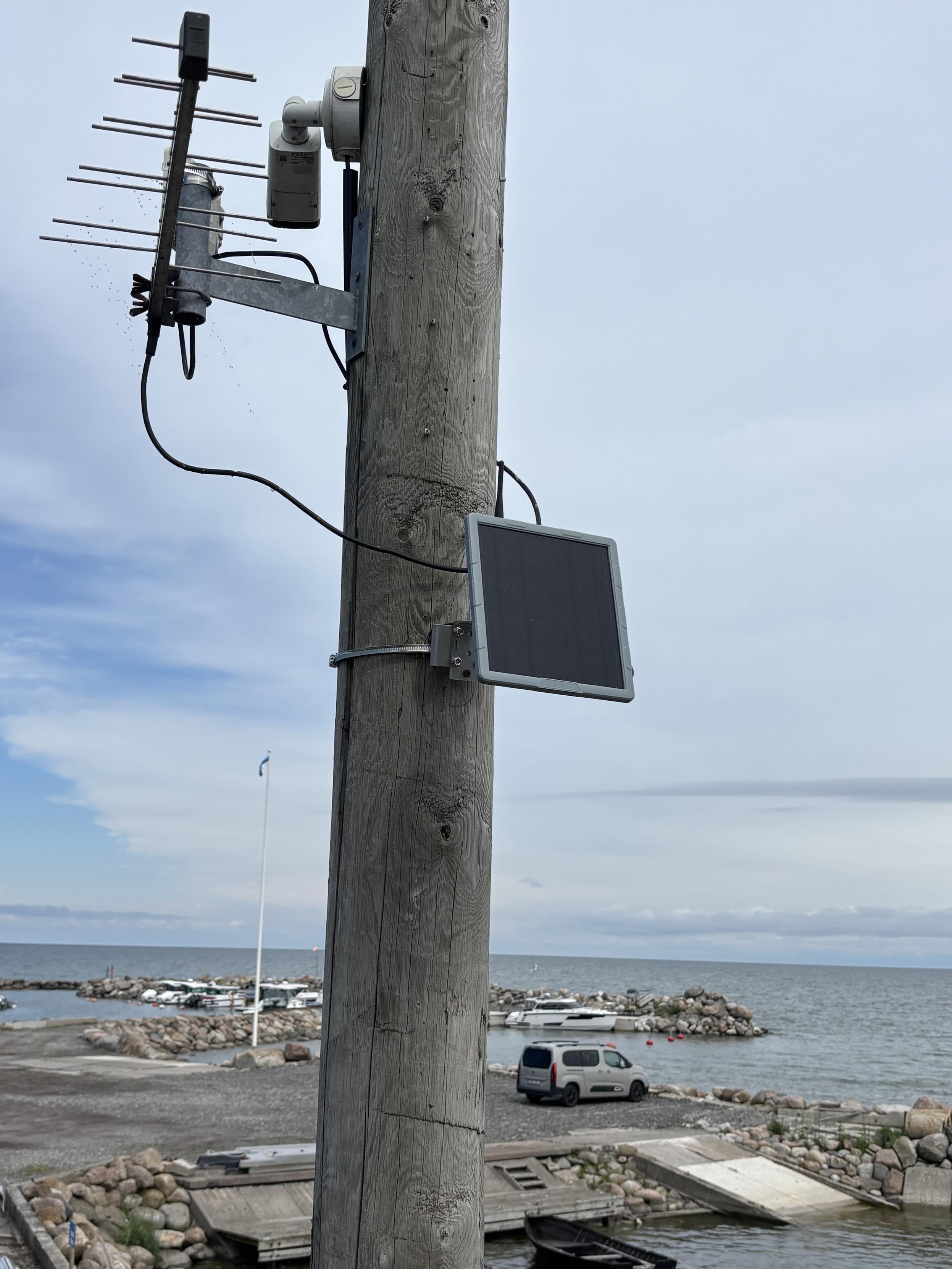 A wooden utility pole with antennas and a solar panel mounted on it. In the background, a marina features several boats and a parking area with a vehicle. The sky is partly cloudy over the waterfront.