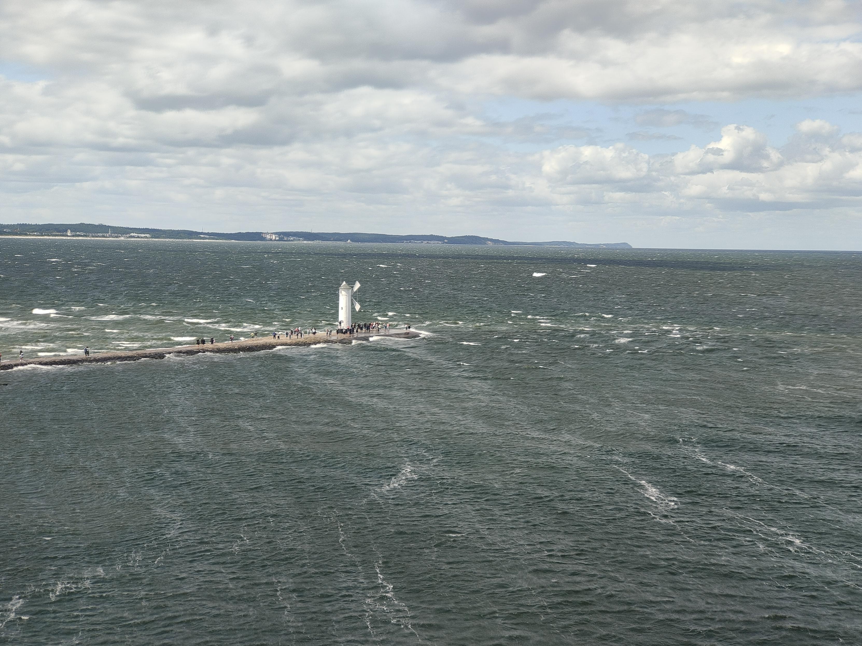 View of open see with windmill at the end of wawe breaker. People standing around look like ants.